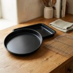 Round and rectangular black non-stick oven trays with a chrome cooling rack on an oak counter — Oven Day bundle
