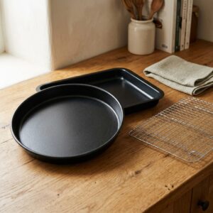Round and rectangular black non-stick oven trays with a chrome cooling rack on an oak counter — Oven Day bundle